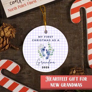 Puede incluir: Adorno navide&ntilde;o redondo con un estampado de cuadros vichy de color lila claro. El adorno tiene el texto "MY FIRST CHRISTMAS AS A Grandma 2025" y presenta huellas de pies azules y vegetaci&oacute;n. Se adjunta una cuerda dorada para colgar.