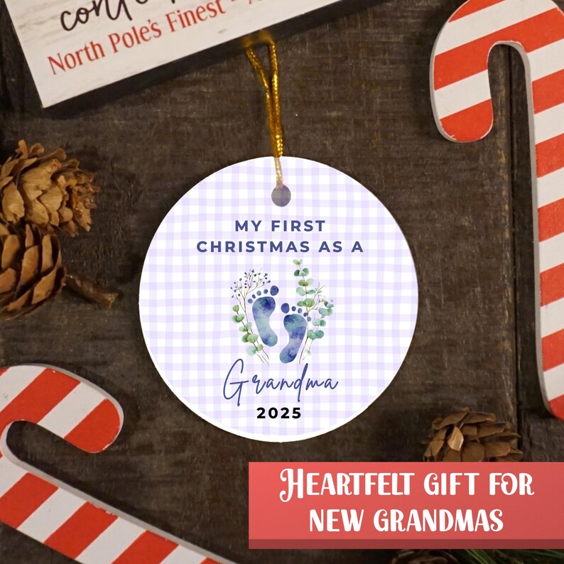 Puede incluir: Adorno navide&ntilde;o redondo con un estampado de cuadros vichy de color lila claro. El adorno tiene el texto "MY FIRST CHRISTMAS AS A Grandma 2025" y presenta huellas de pies azules y vegetaci&oacute;n. Se adjunta una cuerda dorada para colgar.
