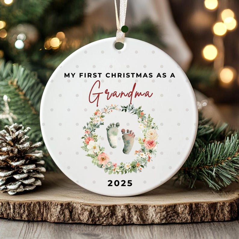 Puede incluir: Adorno navide&ntilde;o redondo de cer&aacute;mica blanca con el texto "MY FIRST CHRISTMAS AS A Grandma". Presenta una corona floral con huellas de beb&eacute; y el a&ntilde;o 2025.