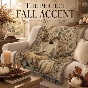 May include: A woven throw blanket featuring a detailed harvest pattern of pumpkins, wheat stalks, and eucalyptus leaves in muted earth tones. The text THE PERFECT FALL ACCENT is printed in dark brown letters above the draped fabric.
