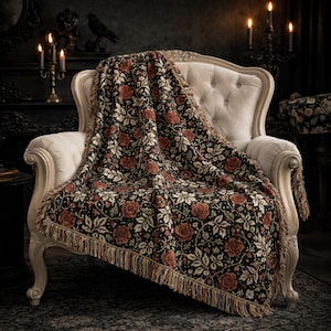 May include: A black throw blanket with a floral pattern of red roses and cream-colored leaves draped over a cream-colored armchair. The blanket has a fringed edge. The background includes candles and a dark, gothic-style interior.