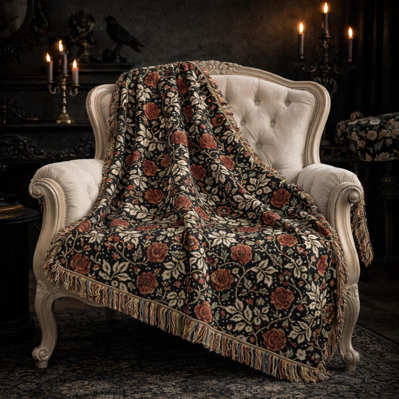 May include: A black throw blanket with a floral pattern of red roses and cream-colored leaves draped over a cream-colored armchair. The blanket has a fringed edge. The background includes candles and a dark, gothic-style interior.
