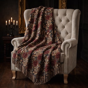 Peut inclure: Une couverture à motifs floraux dans les tons bordeaux, marron et beige, drapée sur un fauteuil beige rembourré. La couverture a un bord à franges et un motif floral détaillé. La chaise a un dossier capitonné et des clous décoratifs.