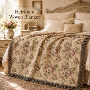 May include: A beige Heirloom Woven Blanket with a floral pattern of pink and green roses. The blanket is draped over a bed with a cream-colored headboard and pillows. The blanket has a fringed edge.