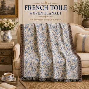 K&ouml;nnte beinhalten: Eine gewebte Decke mit franz&ouml;sischem Toile-de-Jouy-Design, mit blauen Blumen- und Vogelmustern auf cremefarbenem Hintergrund. Die Decke ist &uuml;ber ein Sofa drapiert und hat einen Fransenrand. Der Text "FRENCH TOILE WOVEN BLANKET" ist sichtbar.