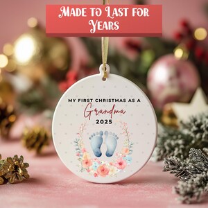 Puede incluir: Adorno navide&ntilde;o redondo de cer&aacute;mica blanca con el texto "MY FIRST CHRISTMAS AS A Grandma 2025". Presenta huellas de pies de beb&eacute; azules y una corona floral. La pancarta superior dice "MADE TO LAST FOR YEARS".