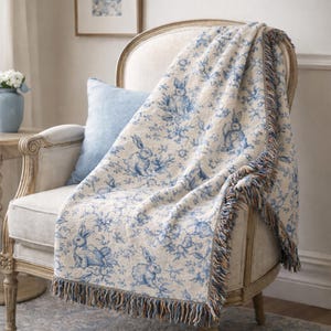 May include: A cream-coloured throw blanket with a blue bunny and floral pattern draped over a chair. The blanket has a fringed edge with blue, brown, and orange threads. A light blue cushion rests on the chair.