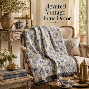 K&ouml;nnte beinhalten: Eine beige Decke mit blauem Blumenmuster, drapiert &uuml;ber einem cremefarbenen Sessel. Die Decke hat einen Fransenrand. Das Bild zeigt auch ein dekoratives Kissen, antike M&ouml;bel und den Text "Elevated Vintage Home Decor."