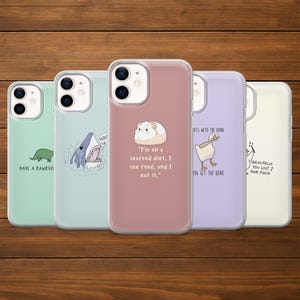 Könnte beinhalten: Eine Sammlung von fünf Handyhüllen mit verschiedenen Designs. Die Hüllen zeigen einen grünen Dinosaurier, einen Hai, ein Meerschweinchen mit Text, eine Gans und Text. Die Hüllen werden auf einer Holzoberfläche präsentiert.