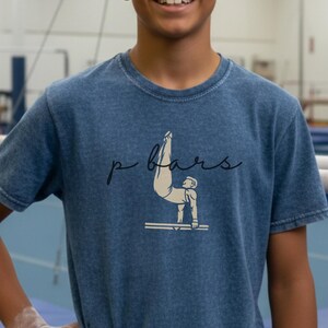 Op de afbeelding: Een blauw t-shirt met de tekst "p bars" in een scriptlettertype en een afbeelding van een gymnast op de brug. De foto is genomen in een gymzaal met brug en andere gymnastiektoestellen op de achtergrond.