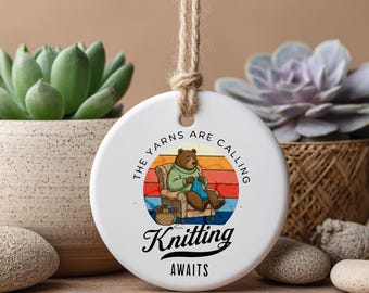 Pode incluir: Enfeite de cerâmica branca com um urso tricotando, o texto "The yarns are calling" e "Knitting Awaits". O urso usa um suéter verde e está sentado em uma cadeira. O fundo tem um design de pôr do sol.