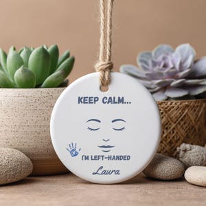 Può includere: Ornamento rotondo in ceramica bianca con un design a faccina sorridente e la scritta "KEEP CALM... I'M LEFT-HANDED Laura". L'ornamento ha un'impronta di mano blu ed è sospeso da un cordoncino di spago naturale. Articolo decorativo.