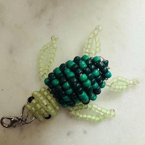 May include: A beaded turtle keyring. The turtle's body is made of dark green and light green beads. The keyring has a silver clasp. The turtle's head and legs are light green.