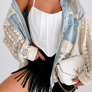 Può includere: Un top a corsetto bianco con spalline sottili e pantaloncini neri con frange. Una giacca di jeans azzurra con maniche in pizzo è indossata sopra. Una borsa bianca trapuntata con catena dorata completa il look.