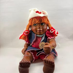 May include: A handcrafted doll with dark skin, red hair in braids, and a white hat with red accents. The doll wears a red outfit with denim accents and holds a small medallion. The doll is sitting with its feet visible.
