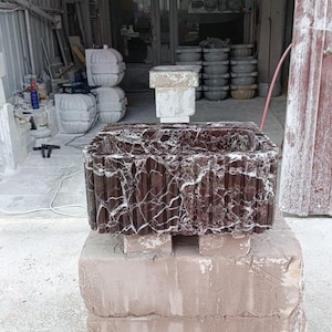 Puede incluir: Un lavabo rectangular de m&aacute;rmol marr&oacute;n oscuro con vetas blancas, apoyado sobre una base de piedra marr&oacute;n claro. El lavabo tiene un exterior texturizado y acanalado. El fondo muestra un taller con otros productos de piedra.