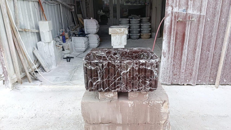 Puede incluir: Un lavabo rectangular de m&aacute;rmol marr&oacute;n oscuro con vetas blancas, apoyado sobre una base de piedra marr&oacute;n claro. El lavabo tiene un exterior texturizado y acanalado. El fondo muestra un taller con otros productos de piedra.