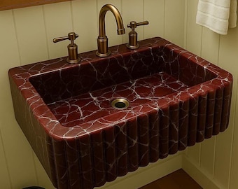 May include: A rectangular wall-mounted bathroom sink featuring a deep red marble finish with white veining. The exterior has a vertical fluted texture. It includes a brass-toned faucet with two matching lever handles and a central drain.