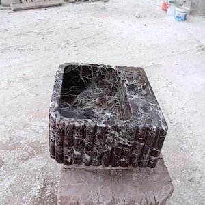 Puede incluir: Un lavabo rectangular de m&aacute;rmol rojo oscuro con vetas blancas. El lavabo tiene una pila y un exterior acanalado. Se exhibe sobre una base de piedra en bruto, mostrando una est&eacute;tica natural y r&uacute;stica. El lavabo es probablemente para un ba&ntilde;o o un espacio exterior.