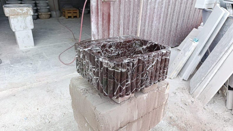 Puede incluir: Un lavabo rectangular de m&aacute;rmol marr&oacute;n oscuro con vetas blancas, apoyado sobre una base de piedra marr&oacute;n claro. El lavabo tiene un exterior texturizado y acanalado. El dise&ntilde;o general sugiere un enfoque en los accesorios de ba&ntilde;o.
