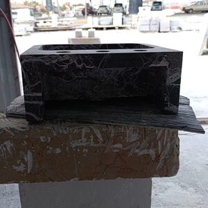 Puede incluir: Un lavabo de ba&ntilde;o de m&aacute;rmol gris oscuro con forma rectangular y tres orificios circulares. El lavabo est&aacute; colocado sobre una base de piedra rugosa de color marr&oacute;n claro. El m&aacute;rmol tiene vetas blancas.