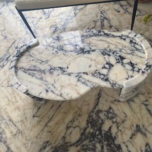 Puede incluir: Una mesa de centro con forma de riñón y tapa de mármol pulido. El mármol presenta un llamativo patrón de vetas blancas, negras y doradas. La mesa tiene bordes redondeados y un diseño moderno, ideal para un salón.