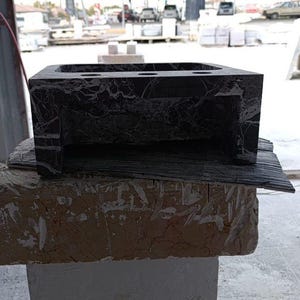 Puede incluir: Un lavabo de ba&ntilde;o de m&aacute;rmol gris oscuro con forma rectangular y un interior ahuecado. El lavabo tiene una superficie texturizada y est&aacute; colocado sobre una base de piedra rugosa de color marr&oacute;n claro. El lavabo tiene tres agujeros circulares.