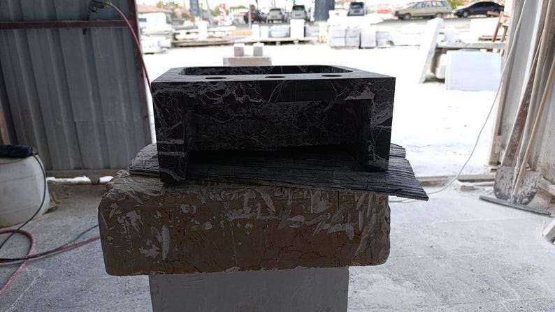 Puede incluir: Un lavabo de ba&ntilde;o de m&aacute;rmol gris oscuro con forma rectangular y un interior ahuecado. El lavabo tiene una superficie texturizada y est&aacute; colocado sobre una base de piedra rugosa de color marr&oacute;n claro. El lavabo tiene tres agujeros circulares.