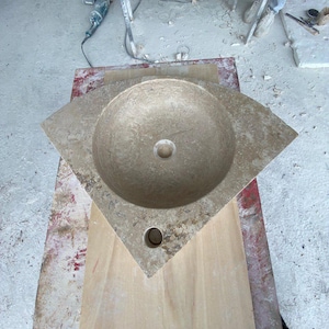 Peut inclure: Un lavabo d'angle triangulaire sculpté dans une pierre naturelle beige à la surface texturée et tachetée. La vasque est circulaire et centrée, avec un trou d'évacuation central et un trou plus petit pour la robinetterie à la pointe avant.