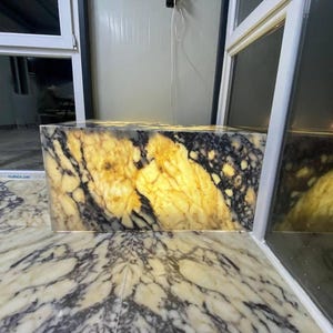 May include: A rectangular onyx block with a striking pattern of black and gold veining. The stone is illuminated from within, creating a warm, glowing effect. The surface below the block has a similar marble pattern.