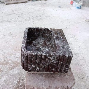 Puede incluir: Un lavabo rectangular de m&aacute;rmol rojo oscuro con vetas blancas. El lavabo tiene una cubeta y un exterior acanalado. Se exhibe sobre una base de piedra en bruto, mostrando una est&eacute;tica natural y r&uacute;stica. El fondo muestra un patio de piedra.