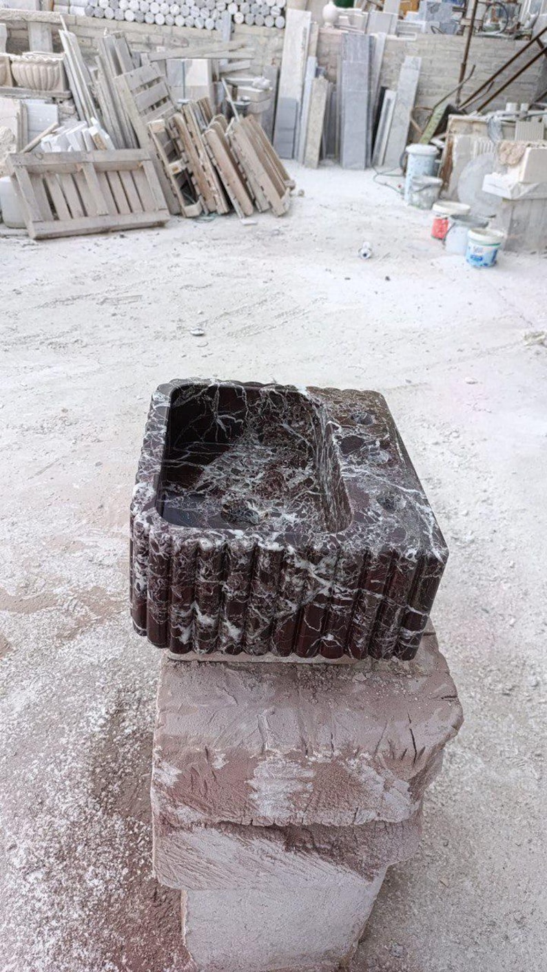 Puede incluir: Un lavabo rectangular de m&aacute;rmol rojo oscuro con vetas blancas. El lavabo tiene una cubeta y un exterior acanalado. Se exhibe sobre una base de piedra en bruto, mostrando una est&eacute;tica natural y r&uacute;stica. El fondo muestra un patio de piedra.