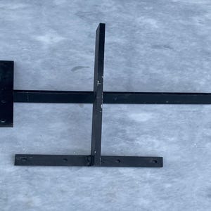 May include: A black metal shelf bracket featuring a T-shaped design with a vertical support bar and a horizontal mounting plate. The bracket includes pre-drilled circular holes along the base and the vertical arm for secure installation.