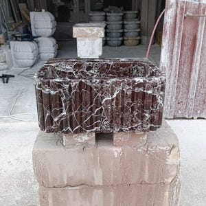Puede incluir: Un lavabo rectangular de piedra marr&oacute;n oscuro con vetas blancas, apoyado sobre una pila de ladrillos. El lavabo tiene un exterior texturizado y acanalado. El fondo muestra un taller con otras piezas de piedra.