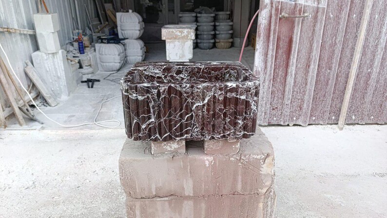 Puede incluir: Un lavabo rectangular de piedra marr&oacute;n oscuro con vetas blancas, apoyado sobre una pila de ladrillos. El lavabo tiene un exterior texturizado y acanalado. El fondo muestra un taller con otras piezas de piedra.