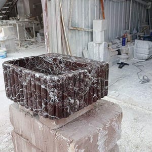 Puede incluir: Un lavabo rectangular de m&aacute;rmol marr&oacute;n oscuro con vetas blancas. El lavabo tiene un dise&ntilde;o acanalado vertical texturizado en el exterior. Se exhibe sobre una base de piedra en bruto en un taller.