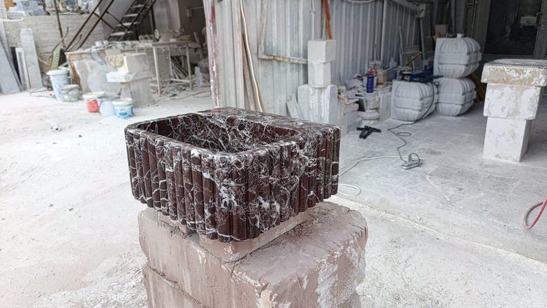 Puede incluir: Un lavabo rectangular de m&aacute;rmol marr&oacute;n oscuro con vetas blancas. El lavabo tiene un dise&ntilde;o acanalado vertical texturizado en el exterior. Se exhibe sobre una base de piedra en bruto en un taller.