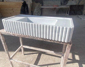 May include: A rectangular, white marble bathtub with a fluted exterior design. The tub is set on a weathered metal stand. The bathroom fixture is in a workshop setting, showcasing the craftsmanship.
