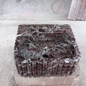 Puede incluir: Un lavabo rectangular de m&aacute;rmol marr&oacute;n oscuro con vetas blancas. El lavabo tiene un borde decorativo acanalado y tres orificios para grifos. El lavabo est&aacute; sobre una superficie gris claro.