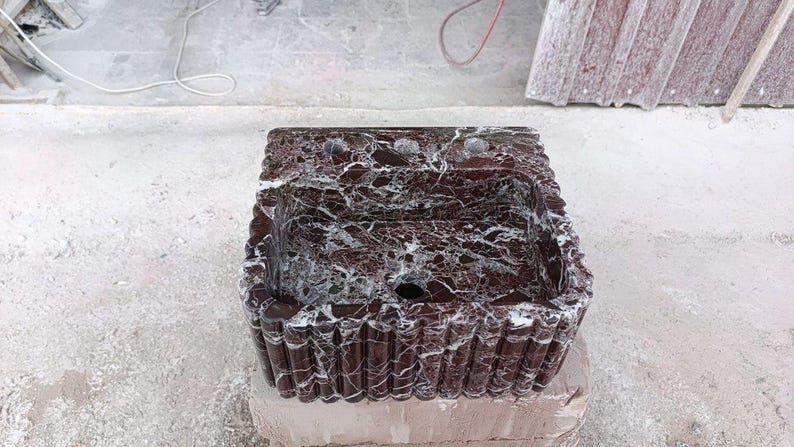 Puede incluir: Un lavabo rectangular de m&aacute;rmol marr&oacute;n oscuro con vetas blancas. El lavabo tiene un borde decorativo acanalado y tres orificios para grifos. El lavabo est&aacute; sobre una superficie gris claro.