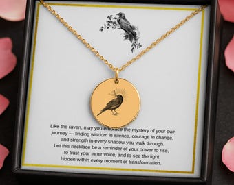 May include: Gold-colored necklace with a circular pendant featuring a raven illustration. The pendant is displayed in a black box with text that reads, "Like the raven, may you embrace the mystery of your own journey..." The necklace is on a white background.