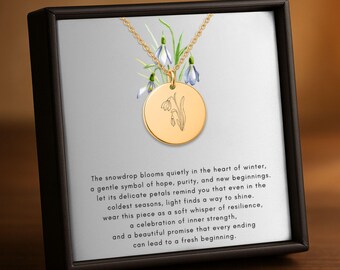 May include: Gold-colored necklace with a round pendant featuring a snowdrop flower design. The necklace is displayed in a black box with a printed background of snowdrop flowers and text. The text reads: "The snowdrop blooms quietly in the heart of winter..."