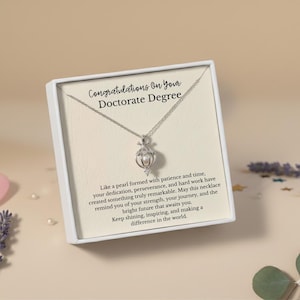 Puede incluir: Un collar plateado con un colgante de perla se muestra en una caja de regalo blanca. La caja tiene el texto "Congratulations On Your Doctorate Degree". El collar está sobre un fondo beige claro.