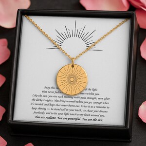 Könnte beinhalten: Eine goldfarbene Halskette mit einem runden Anhänger mit einem Sonnenstrahl-Design. Die Halskette wird in einer schwarzen Schachtel mit weißem Hintergrund und inspirierendem Text präsentiert. Der Anhänger hängt an einer zarten Kette.