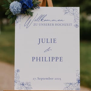 May include: A white wedding welcome sign on a wooden easel, adorned with blue floral accents and a bouquet of blue flowers. The sign reads "Willkommen zu unserer Hochzeit" and lists the names "Julie & Philippe" with the date "27. September 2025".