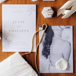 Könnte beinhalten: Hochzeitseinladung und Dankeskarte mit floralen Akzenten. Die Einladung lautet "The Wedding of Julie and Philippe" mit Datum und Ort. Eine kleine Schachtel enthält Ringe. Eine goldene Kette und weißer Stoff sind ebenfalls vorhanden.