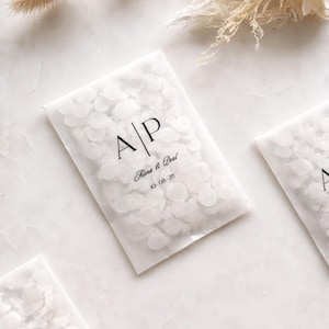 Könnte beinhalten: Weiße Konfetti-Päckchen mit den Initialen "A/P" und den Namen "Alina & Deal" sowie dem Datum "10-08-20" in Schwarz gedruckt. Die Päckchen sind mit weißen Konfetti gefüllt und auf einer weißen Oberfläche mit Trockenblumen angeordnet.