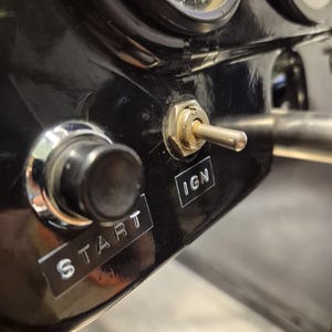 May include: Close-up of a black dashboard with gauges and switches. A toggle switch labeled "IGN" is in the center, with a round button labeled "START" to the left. The gauges have white markings on a black background.