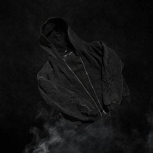 May include: Black zip-up hoodie with a hood and detailed embroidered patterns on the sleeves. The dark garment has a distressed, vintage appearance, set against a smoky, dark background.