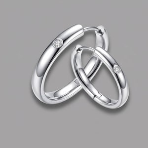 May include: A pair of shiny silver hoop earrings. Each earring features a small, sparkling gemstone. The earrings are displayed against a gray background, highlighting their polished, reflective surface. The design is simple and elegant.